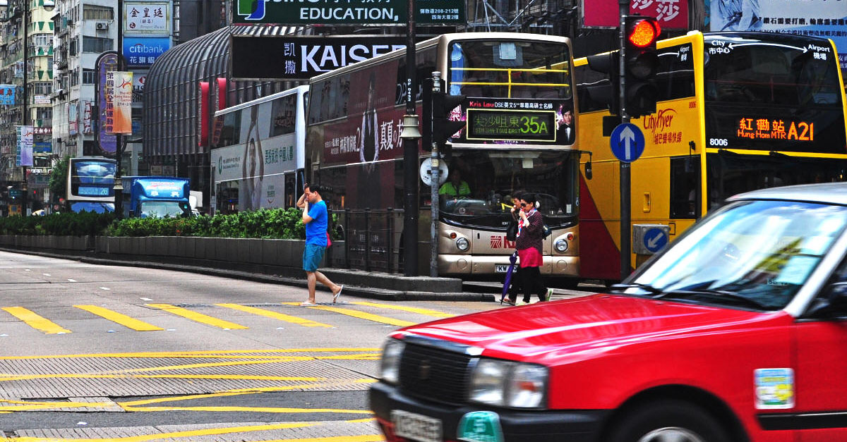 Busy Nathan Road intersection in Hong Kong with double-decker buses, red taxi, and pedestrians crossing amid urban signage
